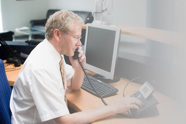 Bild vergrößern: Ein männlicher Mitarbeiter mit Brille, weißem Hemd und Krawatte sitzt an einem Schreibtisch und telefoniert über ein Festnetz-Telefon.
